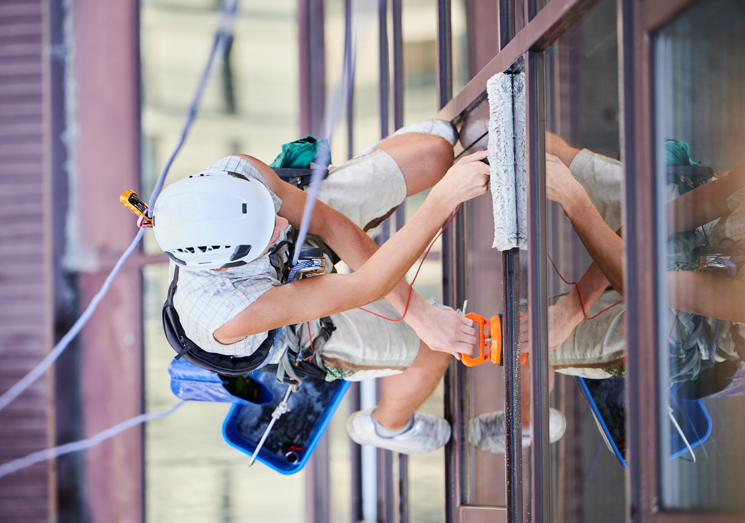 Industrial mountaineering worker washing glass windows of high-rise building, hanging on safety climbing rope. Man window cleaner in protective helmet cleaning skyscraper facade. Top view.