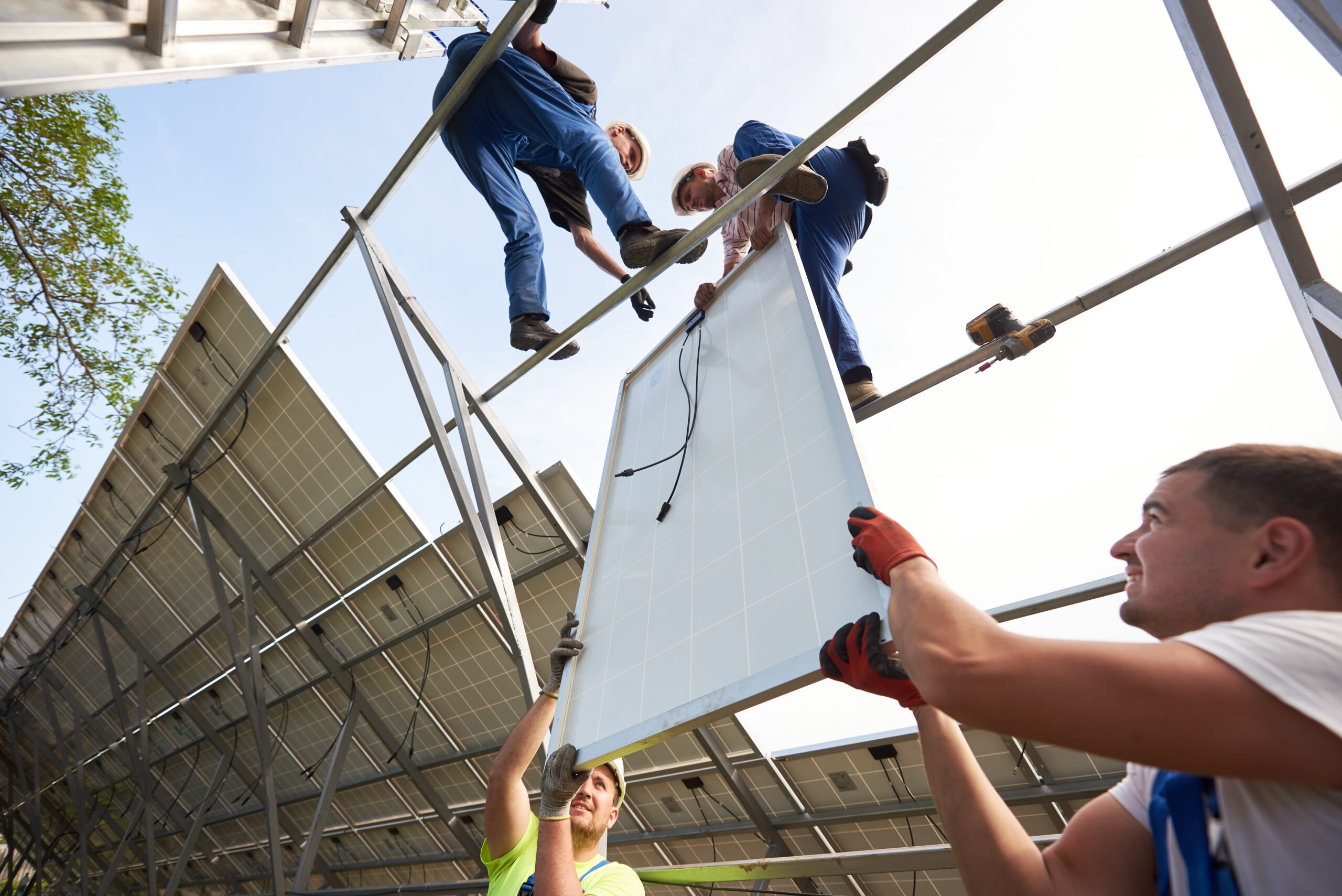 Bottom view from inside of stand-alone exterior solar system with four engineers mounting heavy panel on tall steel platform on bright clear blue sky background. Ecological energy production concept.