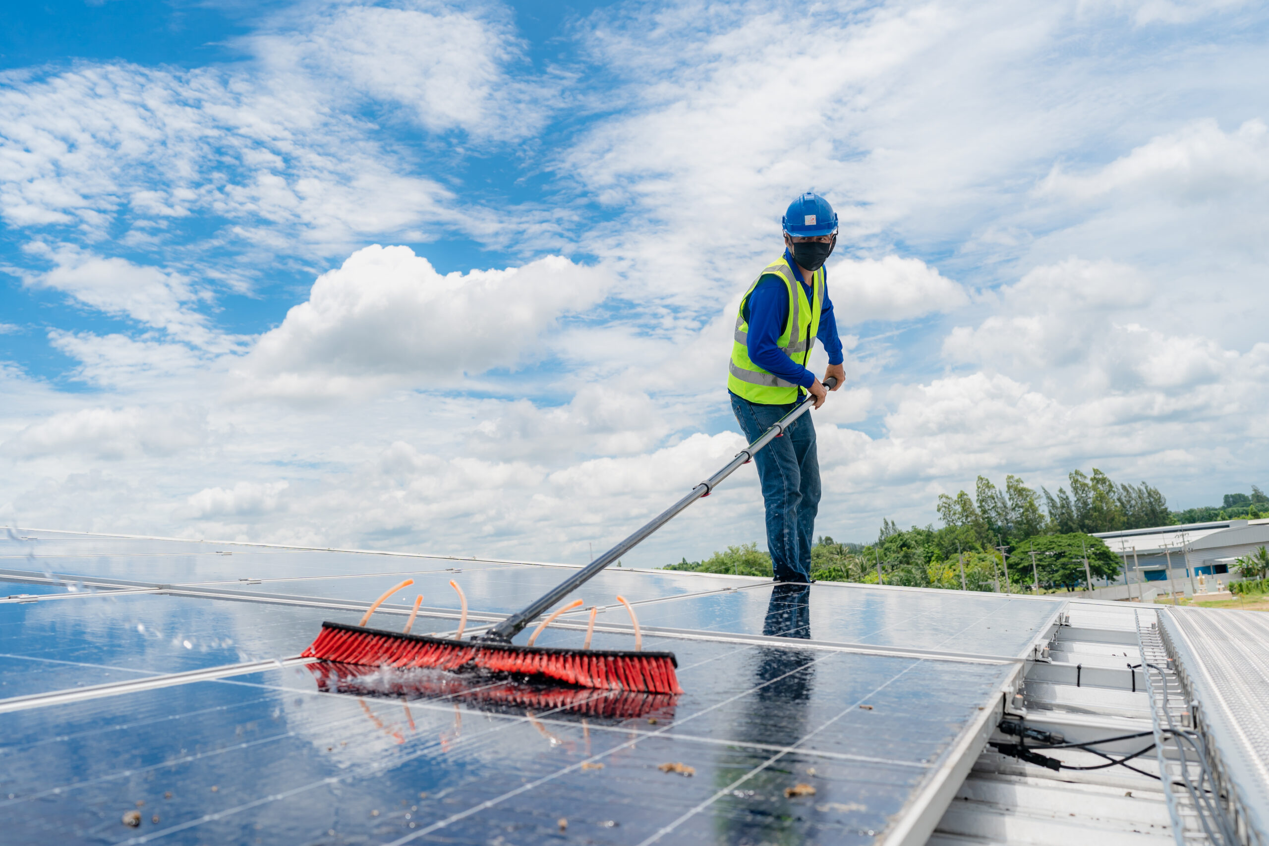 Professional worker cleaning solar panels with brush and washing with water on roof structure of building factory. Technician using mop to clean the dirty and dust, green electricity energy technology