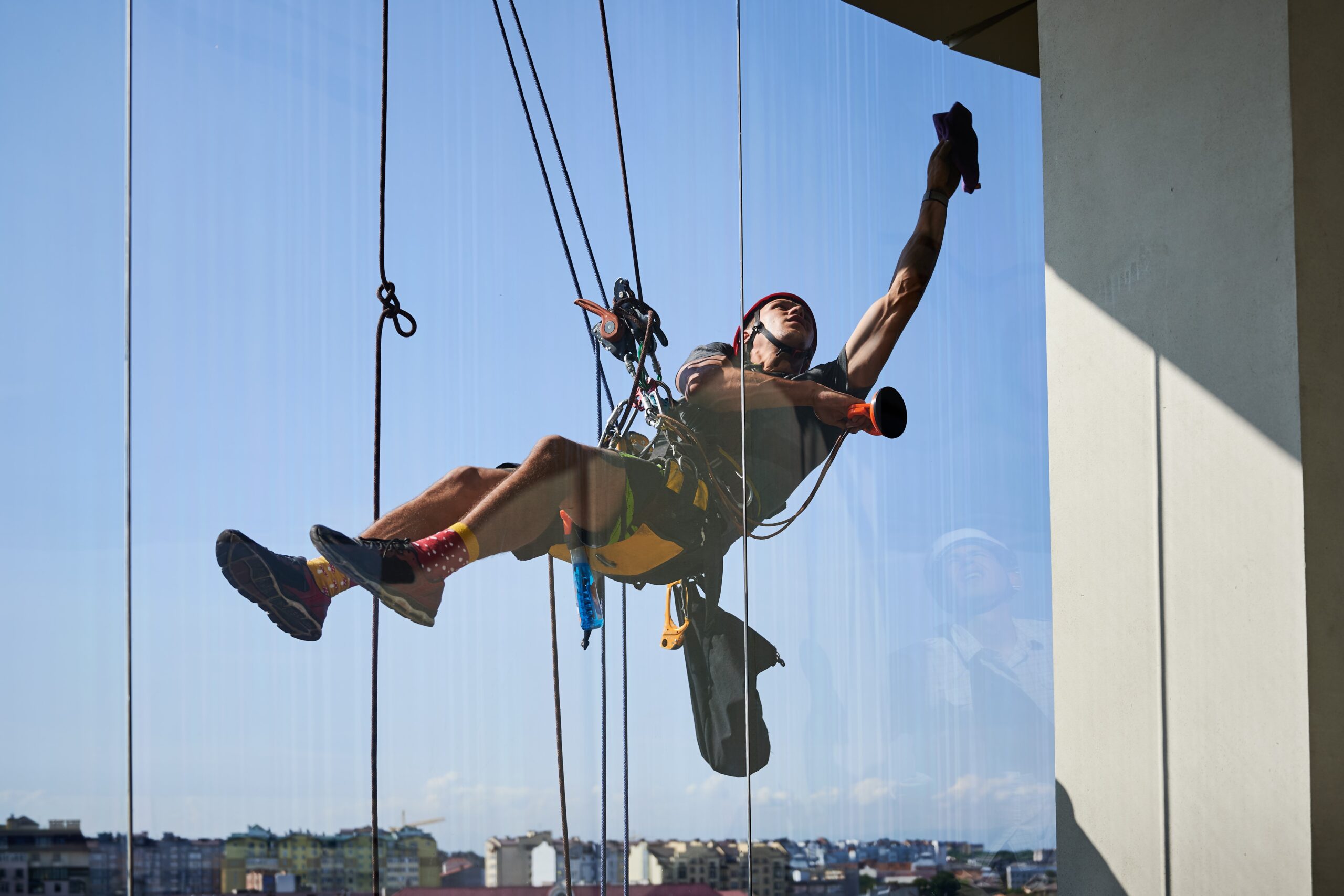 Industrial mountaineering worker hanging on climbing rope and cleaning window of high-rise building. Man using safety lifting equipment while wiping skyscraper window with rag.