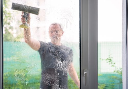 Young man cleans the windows in the house. He splashes detergent, rubs and removes water at the glass. High quality photo