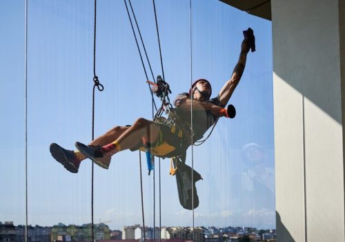 Industrial mountaineering worker hanging on climbing rope and cleaning window of high-rise building. Man using safety lifting equipment while wiping skyscraper window with rag.